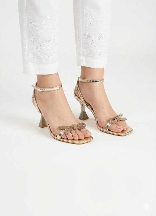 Gold high-heeled sandals with bow details worn with white pants on a light background